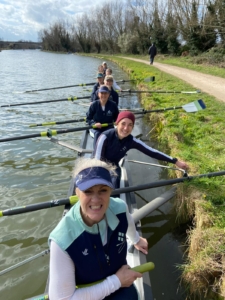Smiling rowers in the Ely women's 1st eight, leaning on the river bank waiting to race.